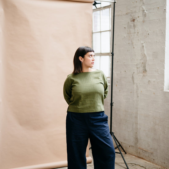 Woman in a green sweater and blue pants standing in a studio with a beige backdrop.