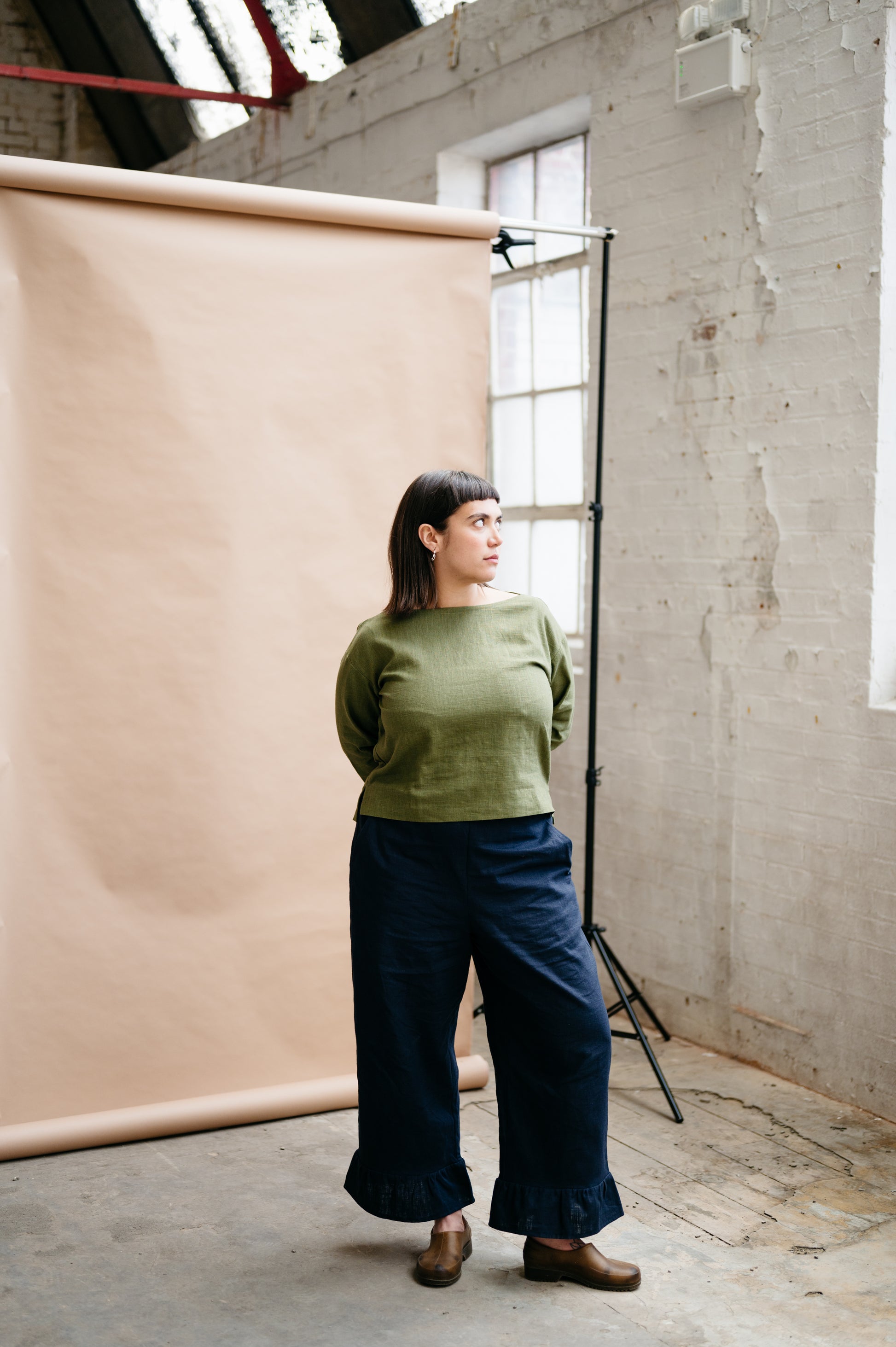 Woman in a green sweater and blue pants standing in a studio with a beige backdrop.