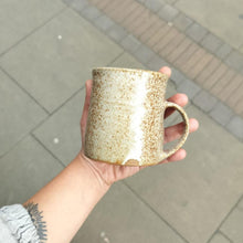 Load image into Gallery viewer, A hand holding a beige ceramic tall mug with brown speckle glaze.
