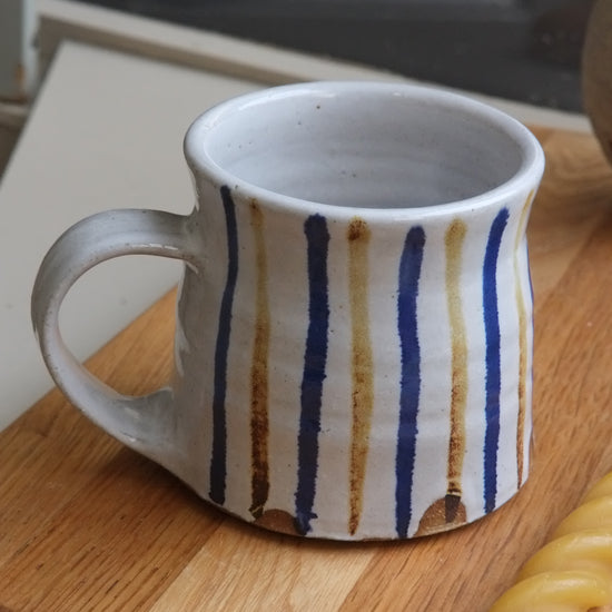 A handmade ceramic mug with a white glaze, decorated with vertical hand-painted stripes in navy blue and warm brown, resting on a wooden cutting board.