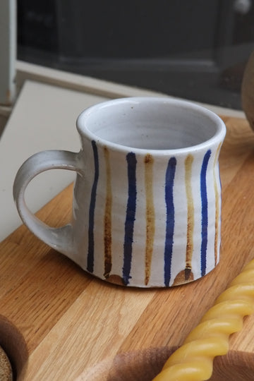 A handmade ceramic mug with a white glaze, decorated with vertical hand-painted stripes in navy blue and warm brown, resting on a wooden cutting board.