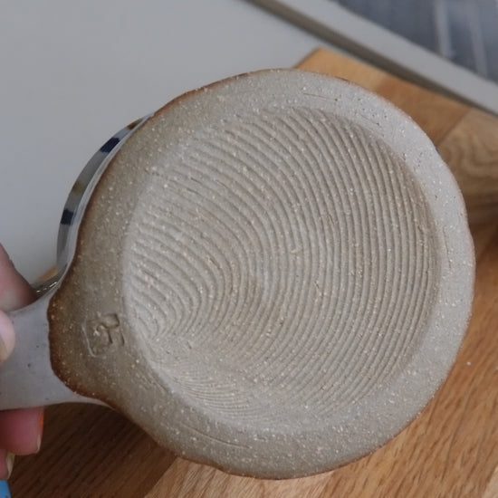 A close-up of a handmade ceramic mug's textured bottom, showing a hand holding it over a wooden surface.