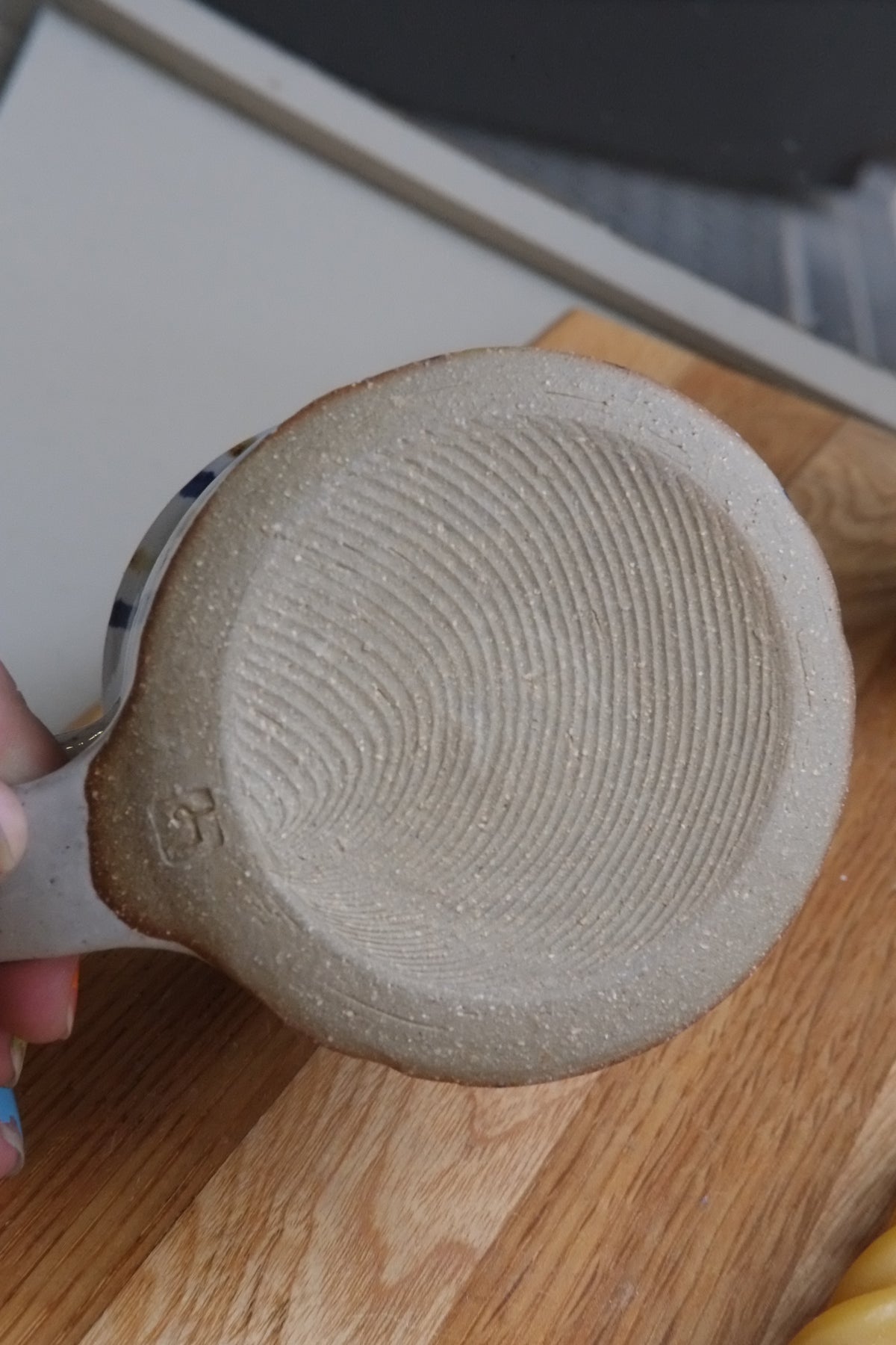A close-up of a handmade ceramic mug's textured bottom, showing a hand holding it over a wooden surface.