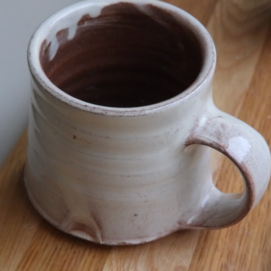 A handmade ceramic mug with a rustic handle, featuring a light beige exterior and a terracotta interior, resting on a wooden cutting board.