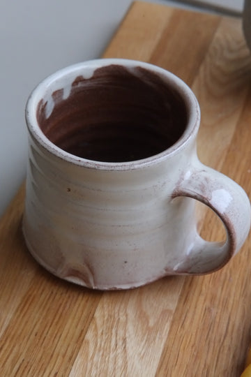 A handmade ceramic mug with a rustic handle, featuring a light beige exterior and a terracotta interior, resting on a wooden cutting board.