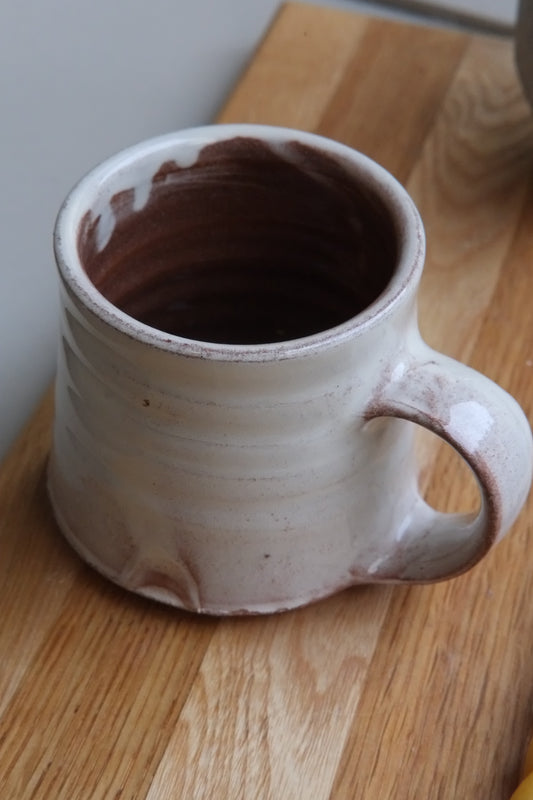 A handmade ceramic mug with a rustic handle, featuring a light beige exterior and a terracotta interior, resting on a wooden cutting board.
