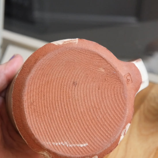 A close-up of a hand holding the bottom of a terracotta-colored ceramic mug or cup, showing the textured circular base and a small white mark, with a wooden table and dark background behind it.