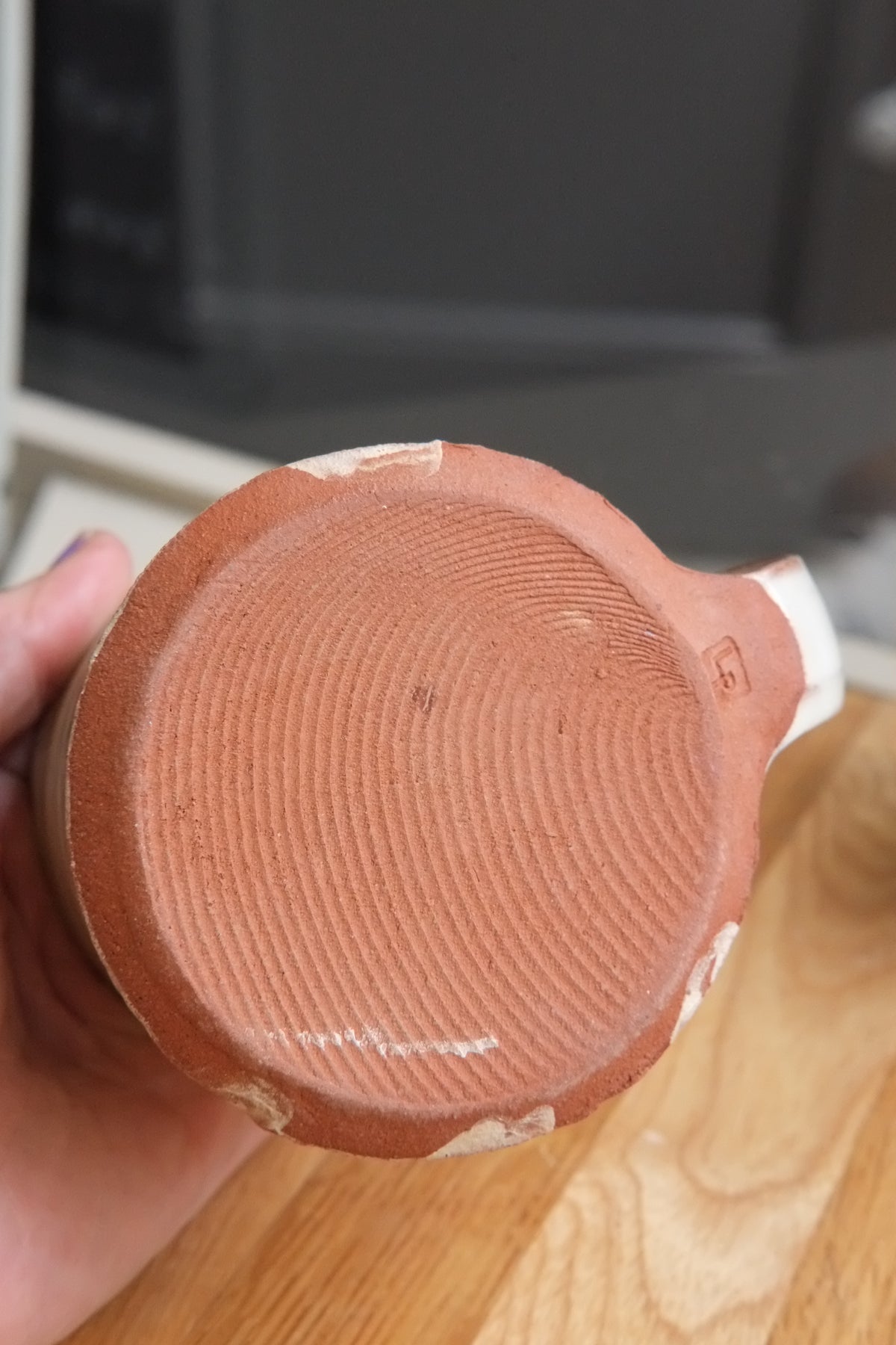 A close-up of a hand holding the bottom of a terracotta-colored ceramic mug or cup, showing the textured circular base and a small white mark, with a wooden table and dark background behind it.