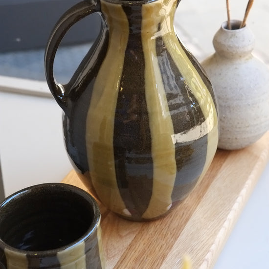 Green stripe ceramic jug with a rounded body and spout on a wooden board surrounded by other ceramics