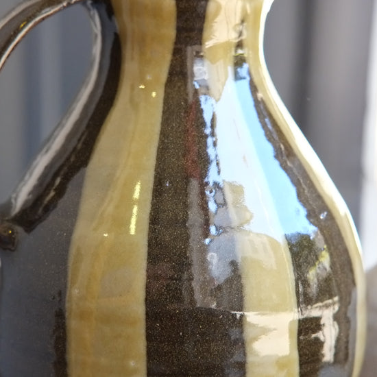 A close-up of a handmade ceramic pitcher with a glossy finish, featuring vertical stripes of dark brown and green