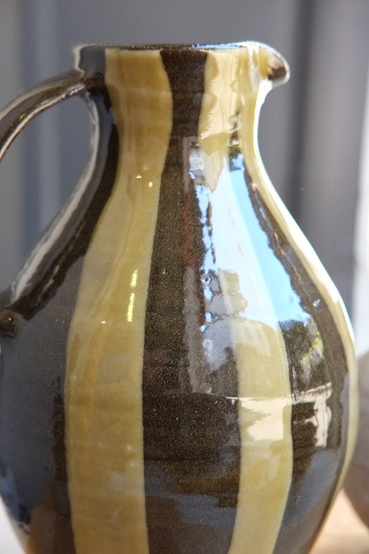A close-up of a handmade ceramic pitcher with a glossy finish, featuring vertical stripes of dark brown and green