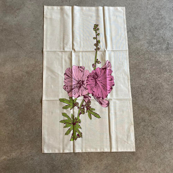A beige cotton tea towel with a floral hollyhock design printed on it, laid out flat.