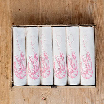 A set of six folded cloth napkins with a pink prawn design, placed in a cardboard box on a wooden surface.