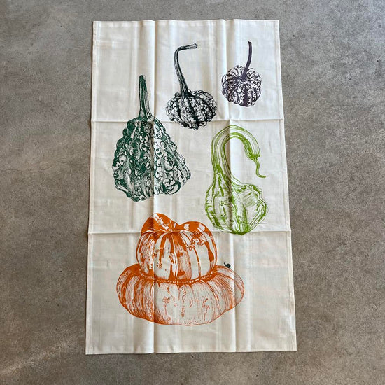 A kitchen towel with a variety of squash and root vegetable illustrations in a norfolk blue, green, and orange color palette on a white background.