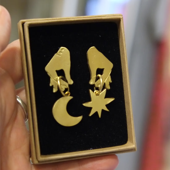 A hand holding a small square gift box with a black velvet insert displaying a pair of gold dangling earrings shaped like hands with a crescent moon and star.