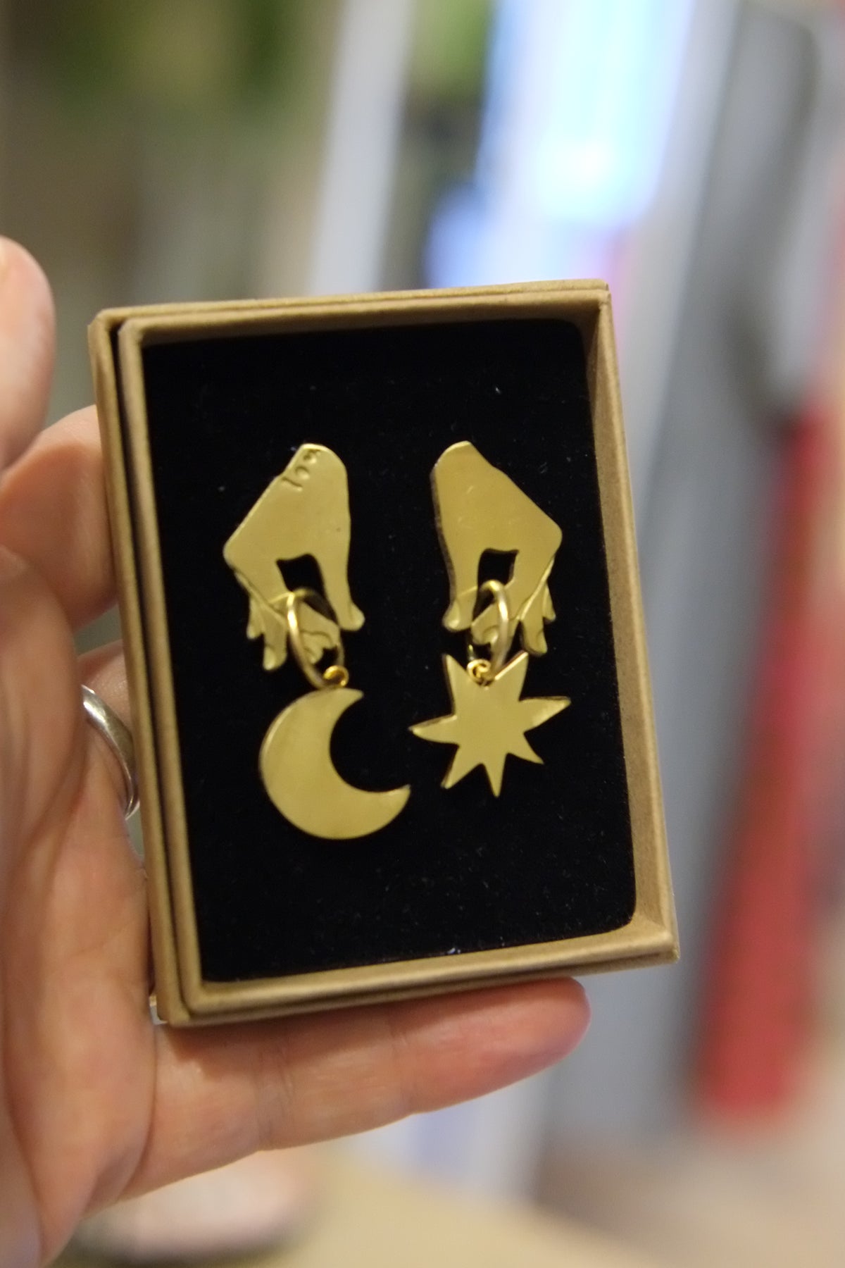 A hand holding a small square gift box with a black velvet insert displaying a pair of gold dangling earrings shaped like hands with a crescent moon and star.