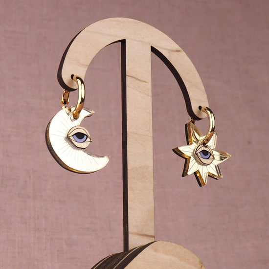 Pair of gold moon and star earrings with eyes on a wooden stand with a pink background