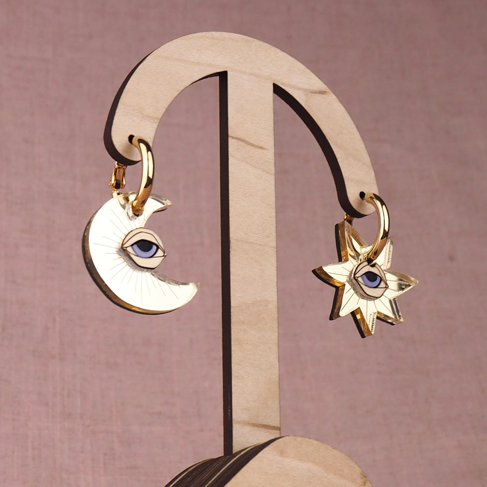 Pair of gold moon and star earrings with eyes on a wooden stand with a pink background
