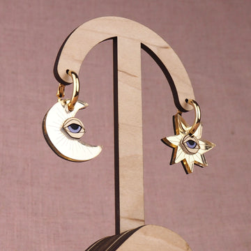 Pair of gold moon and star earrings with eyes on a wooden stand with a pink background