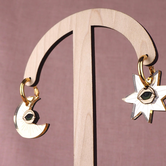 Gold earrings with star and moon designs with eyes on a wooden stand against a pink background