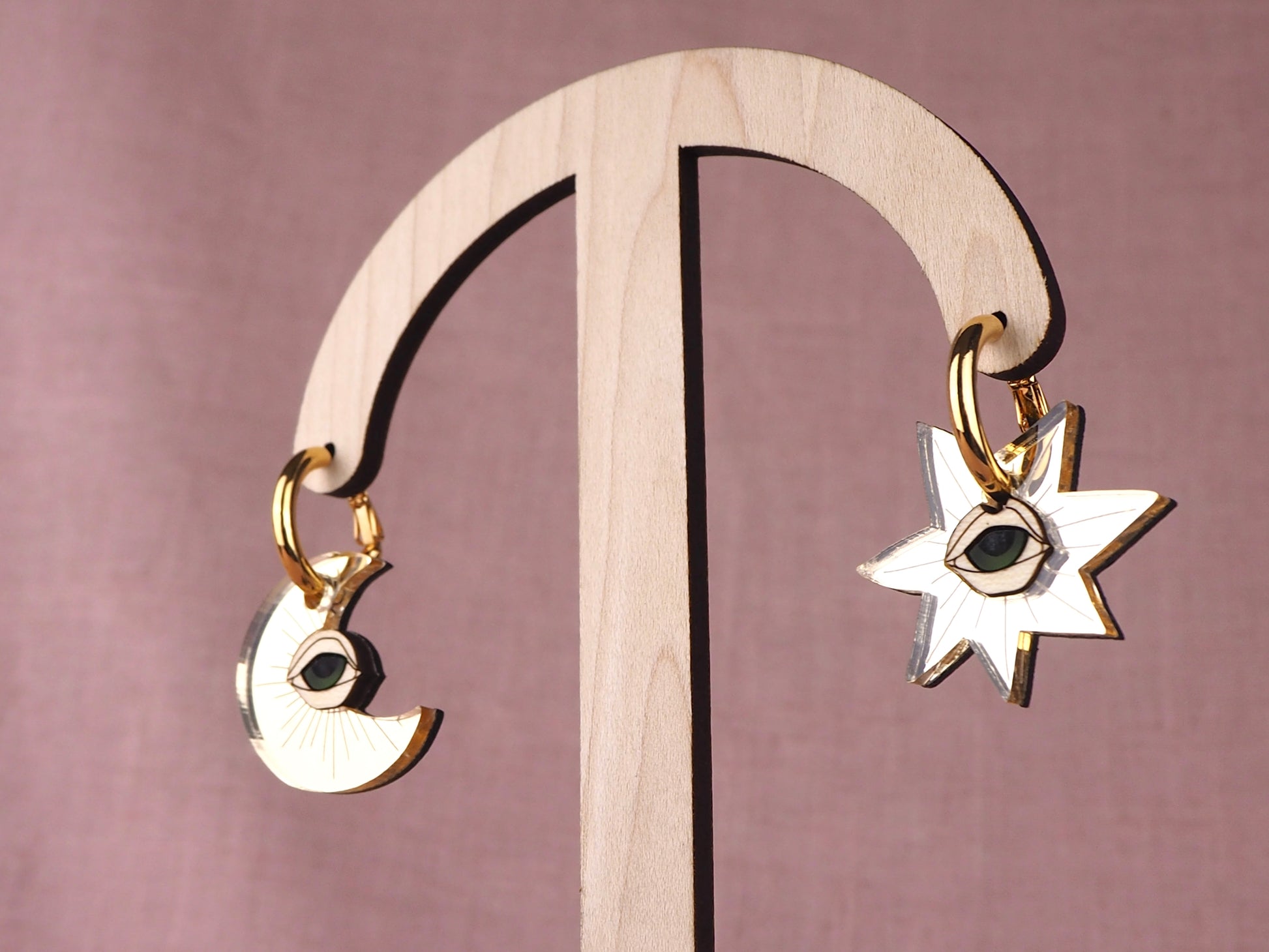 Gold earrings with star and moon designs with eyes on a wooden stand against a pink background