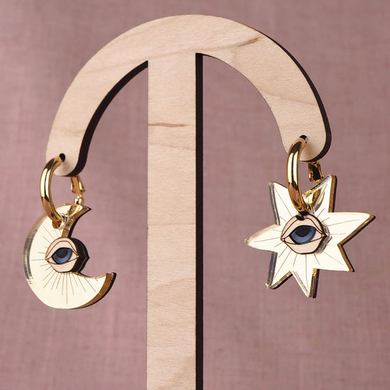Gold earrings with star and crescent moon designs with eyes on a wooden stand against a pink background