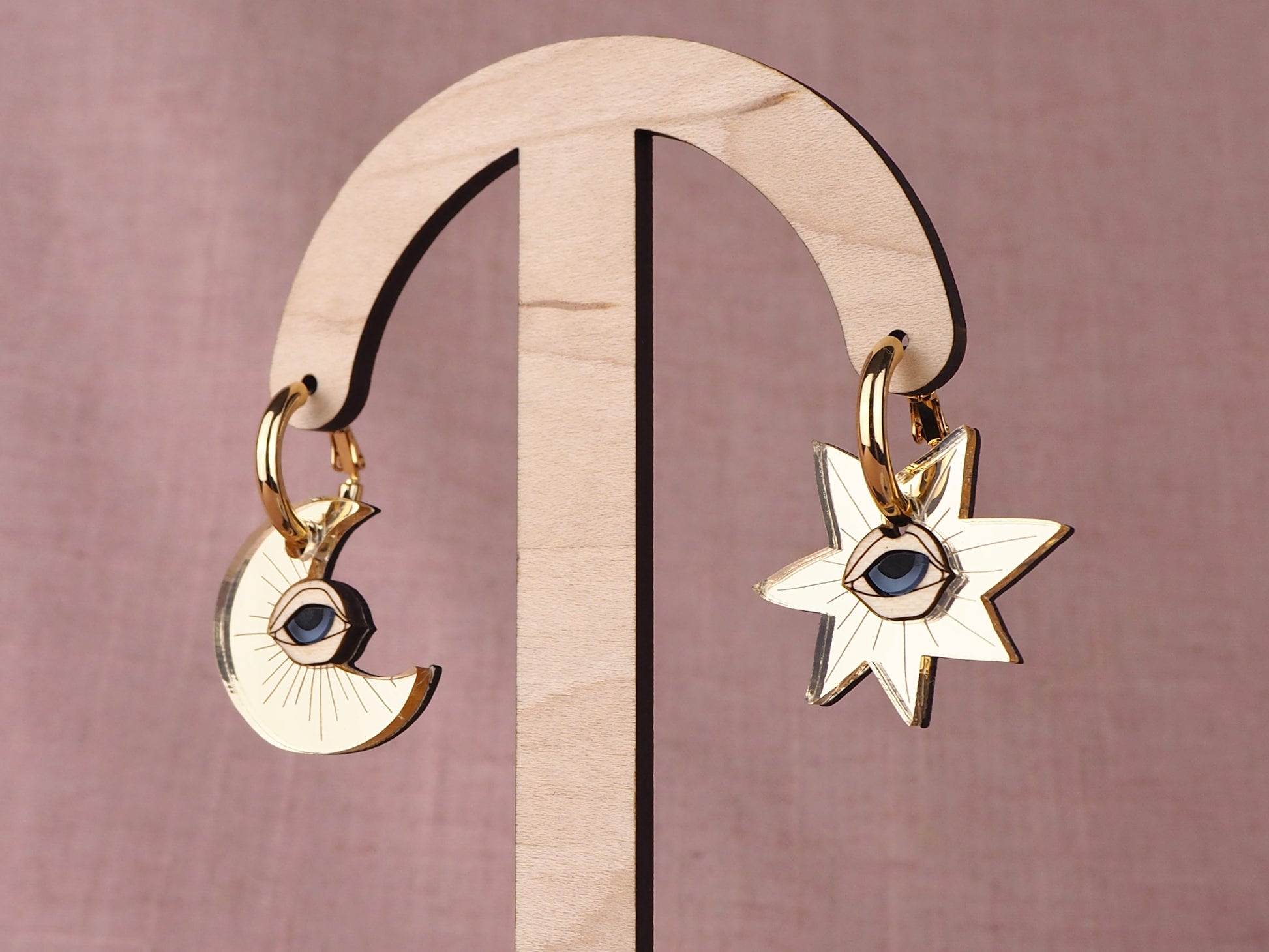 Gold earrings with star and crescent moon designs with eyes on a wooden stand against a pink background