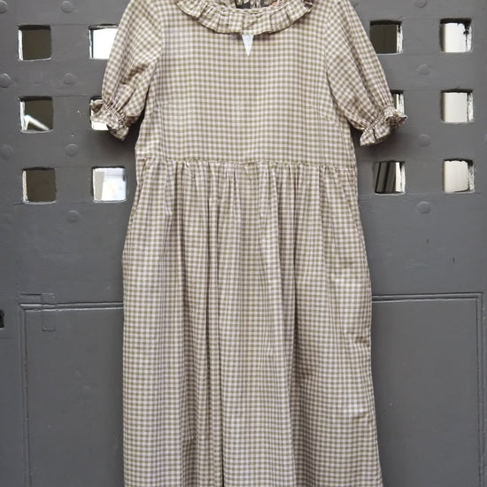 A short-sleeve gingham dress in green and pink, hanging on a wooden hanger against a dark grey perforated metal wall.