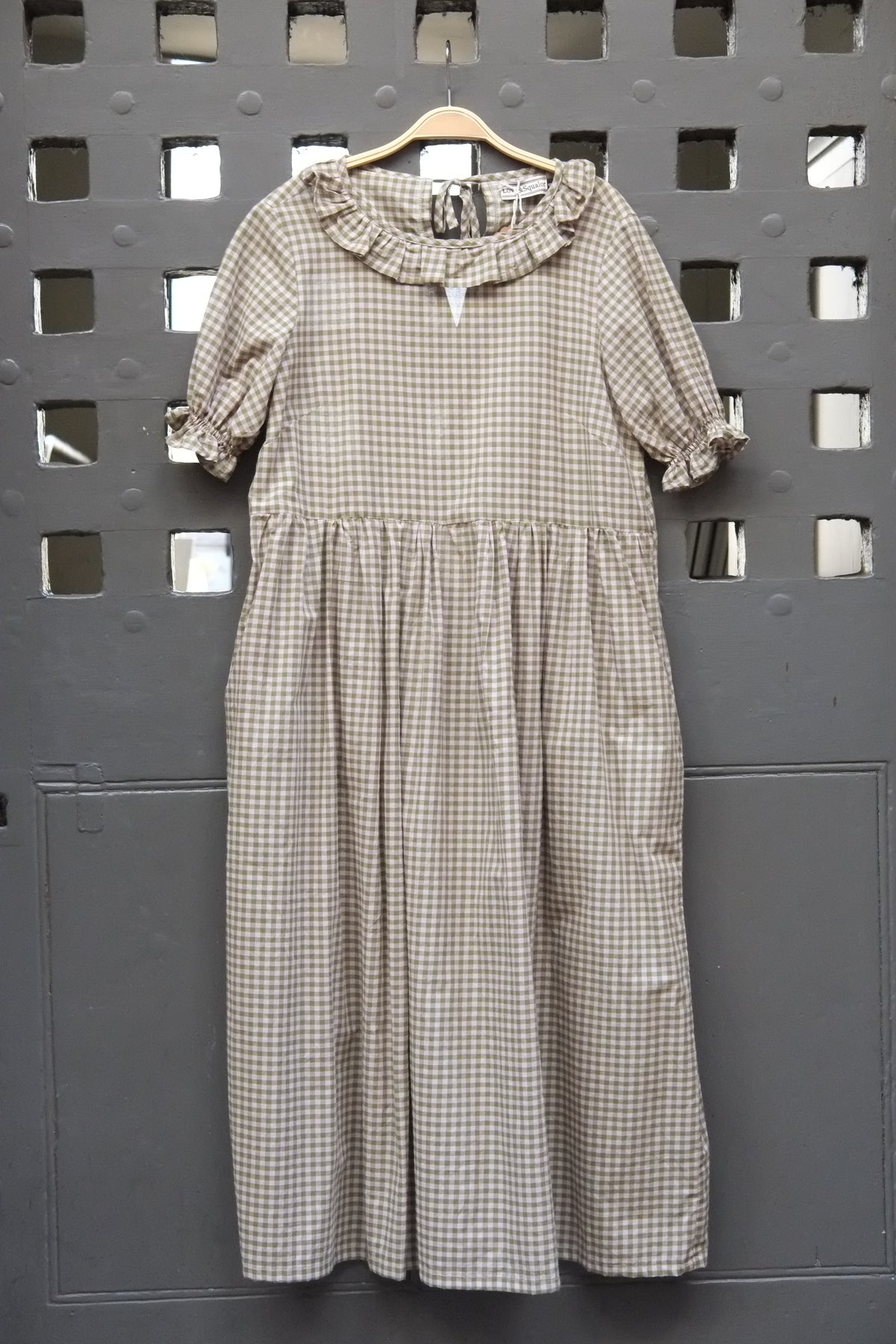 A short-sleeve gingham dress in green and pink, hanging on a wooden hanger against a dark grey perforated metal wall.