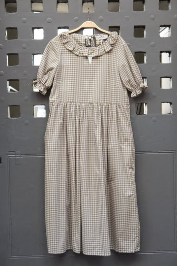 A short-sleeve gingham dress in green and pink, hanging on a wooden hanger against a dark grey perforated metal wall.