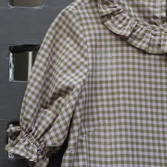 A short-sleeve gingham dress in green and pink, featuring a ruffled collar and gathered elbow-length sleeves, hanging on a wooden hanger against a dark grey panelled door.