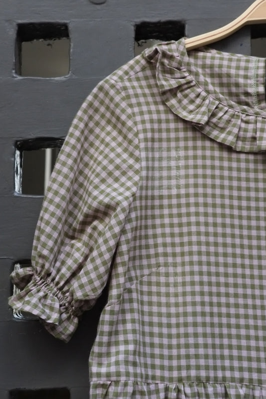 A short-sleeve gingham dress in green and pink, featuring a ruffled collar and gathered elbow-length sleeves, hanging on a wooden hanger against a dark grey panelled door.
