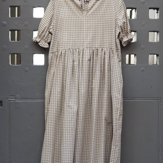 A short-sleeve gingham dress in green and pink hangs on a wooden hanger against a dark grey metal door with square cutouts.