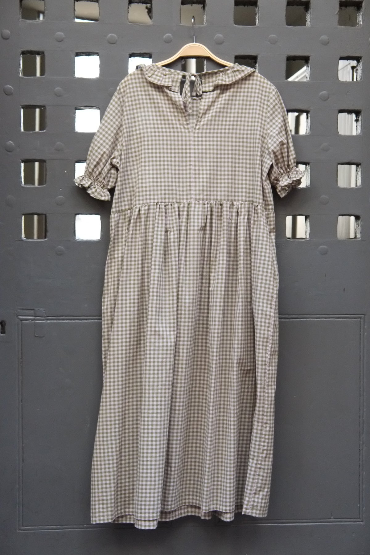 A short-sleeve gingham dress in green and pink hangs on a wooden hanger against a dark grey metal door with square cutouts.
