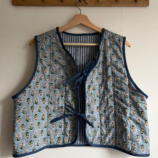 A sleeveless quilted vest with a blue floral print and navy trim, hanging on a wooden hanger from a wooden wall rack.