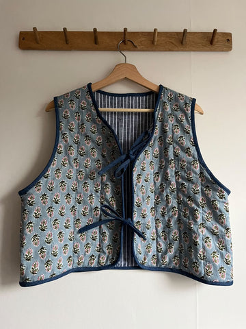 A sleeveless quilted vest with a blue floral print and navy trim, hanging on a wooden hanger from a wooden wall rack.