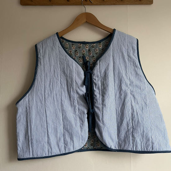 A light blue and white stripe padded quilted vest with navy blue trim and a tie-front closure, hanging on a wooden hanger from a wooden peg rack against a beige wall.