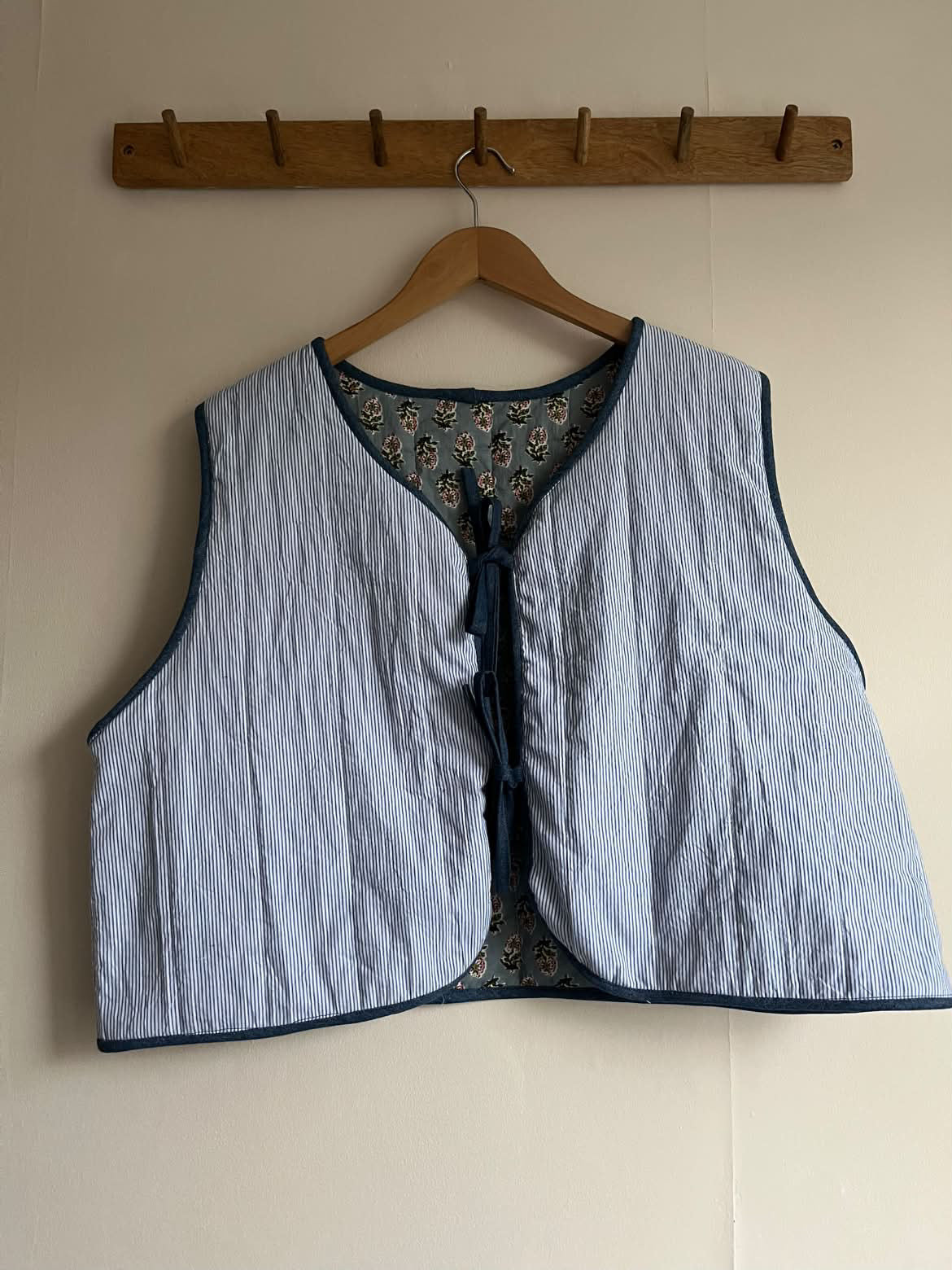 A light blue and white stripe padded quilted vest with navy blue trim and a tie-front closure, hanging on a wooden hanger from a wooden peg rack against a beige wall.