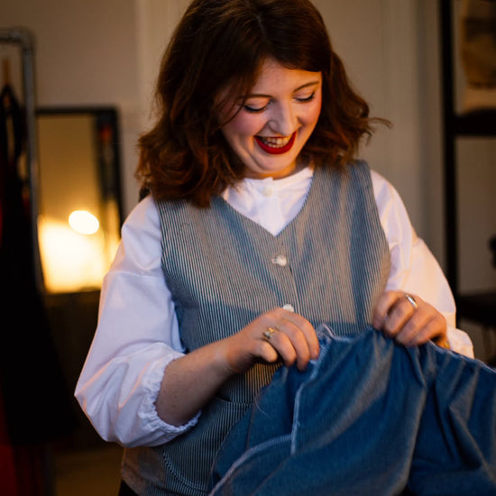 A woman with brown hair smiles while holding up a blue denim garment wearing a blue and white stripe button down waistcoat