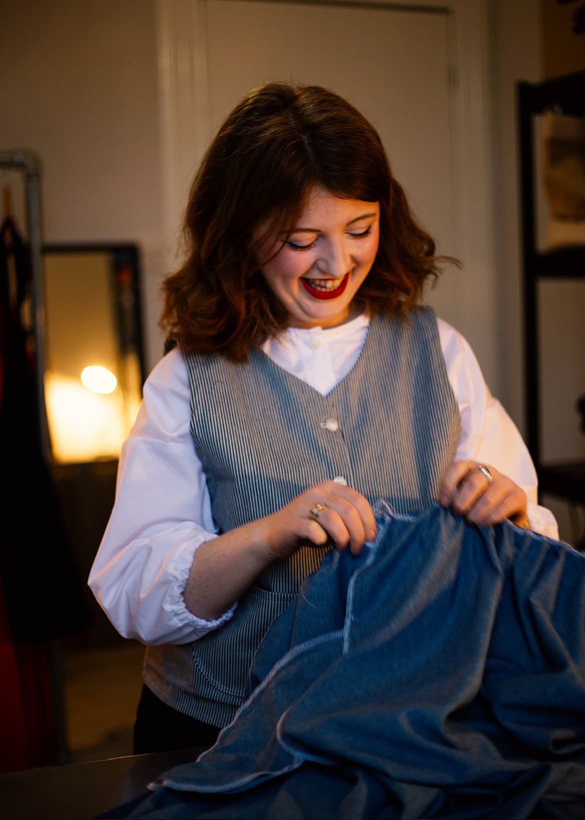 A woman with brown hair smiles while holding up a blue denim garment wearing a blue and white stripe button down waistcoat