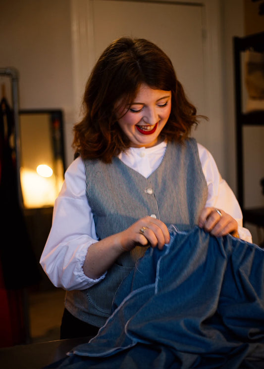 A woman with brown hair smiles while holding up a blue denim garment wearing a blue and white stripe button down waistcoat