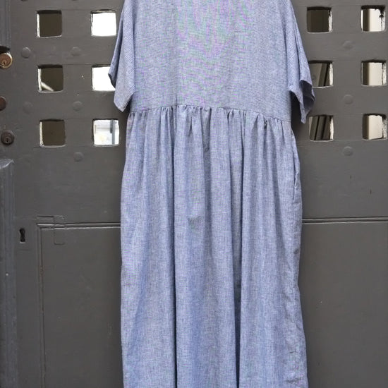 A light blue and white dress with a gathered waist and short sleeves, hanging on a wooden hanger against a dark grey metal door with a grid pattern.