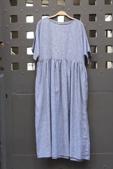 A light blue and white dress with a gathered waist and short sleeves, hanging on a wooden hanger against a dark grey metal door with a grid pattern.