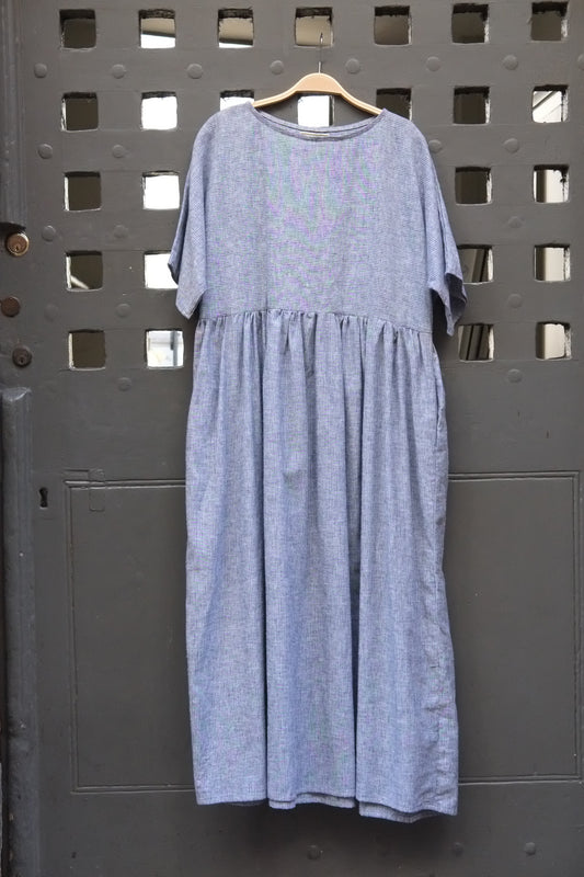 A light blue and white dress with a gathered waist and short sleeves, hanging on a wooden hanger against a dark grey metal door with a grid pattern.