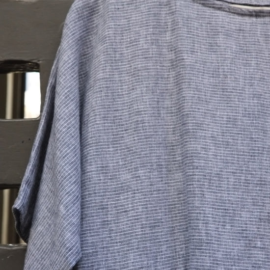 A textured blue and white dress with short wide sleeves and a gathered waist, hanging on a wooden hanger against a dark grey wooden lattice background.