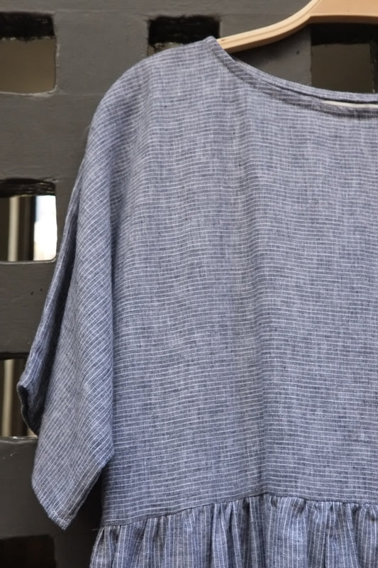 A textured blue and white dress with short wide sleeves and a gathered waist, hanging on a wooden hanger against a dark grey wooden lattice background.