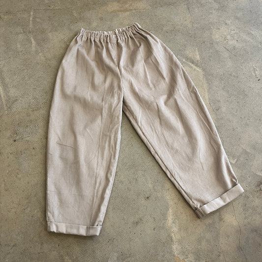 A pair of beige corduroy trousers laid flat on a concrete surface, showing an elastic waistband and a relaxed fit.