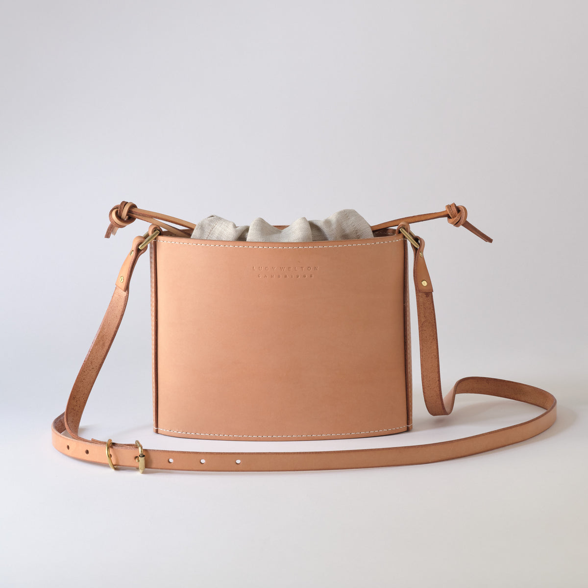 A light tan leather bucket bag with a drawstring top, a matching adjustable shoulder strap, and a light gray fabric dust bag, photographed on a seamless light gray background.