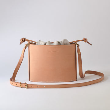 A light tan leather bucket bag with a drawstring top, a matching adjustable shoulder strap, and a light gray fabric dust bag, photographed on a seamless light gray background.