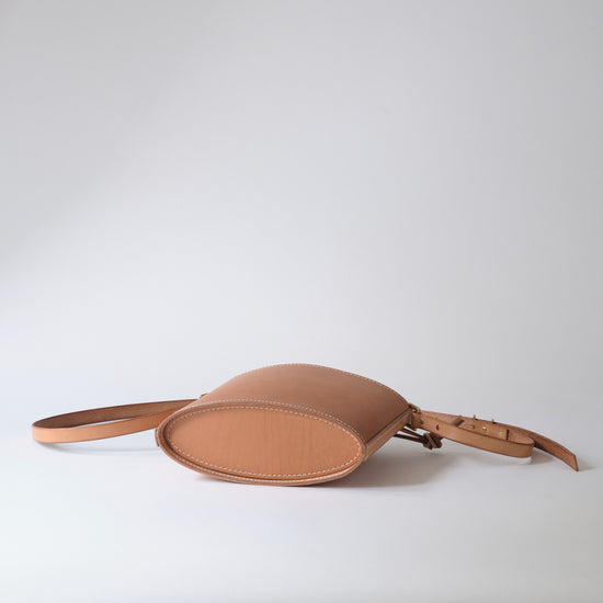 A light tan leather crossbody bag with an oval body, a matching leather strap, and a small leather detail on the side, photographed on a light gray studio background.